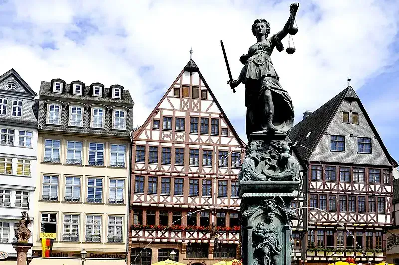 Ein historischer Platz mit traditionellen Fachwerkhäusern und der Gerechtigkeitsbrunnen-Statue der Justitia, die im Vordergrund Waage und Schwert hält. Der Himmel ist teilweise bewölkt und die Gebäude zeichnen sich durch aufwendige Holzarbeiten und malerische Architektur aus.