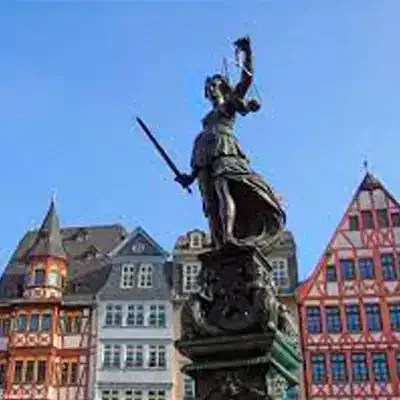 Eine bronzene Statue einer Frau mit Schwert und Waage steht prominent vor einer Reihe farbenfroher, traditioneller europäischer Gebäude mit Giebeldächern. Der Himmel ist klar und blau und zeigt auf diesem historischen Platz eine der Sehenswürdigkeiten der Stadt, die man unbedingt besuchen muss.