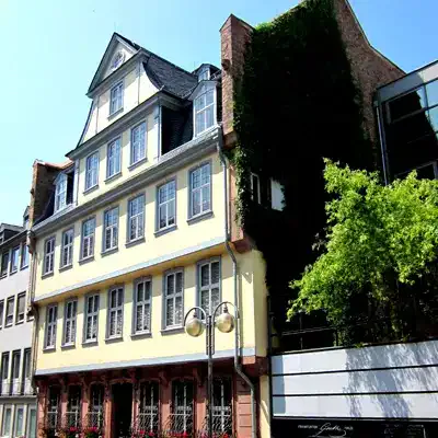 Ein mehrstöckiges Gebäude mit weißen und gelben Außenwänden und zahlreichen Fenstern, gelegen an einer sonnigen Straße. Ein Teil des Gebäudes ist mit grünem Efeu bewachsen. Rechts daneben steht ein modernes Gebäude mit reflektierenden Glaspaneelen, wodurch diese Ecke eine Mischung aus historischem Charme und zeitgenössischen Sehenswürdigkeiten darstellt.