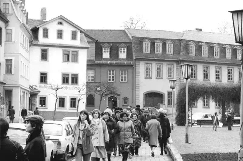 Ein Schwarzweißfoto zeigt eine Straßenszene mit Menschen, die auf einem Bürgersteig vor Gebäuden im traditionellen europäischen Stil spazieren. Die meisten Menschen tragen Winterkleidung. Straßenlaternen säumen den Weg, Autos parken am Rand und im Hintergrund ragt das Goethe-Haus hervor.