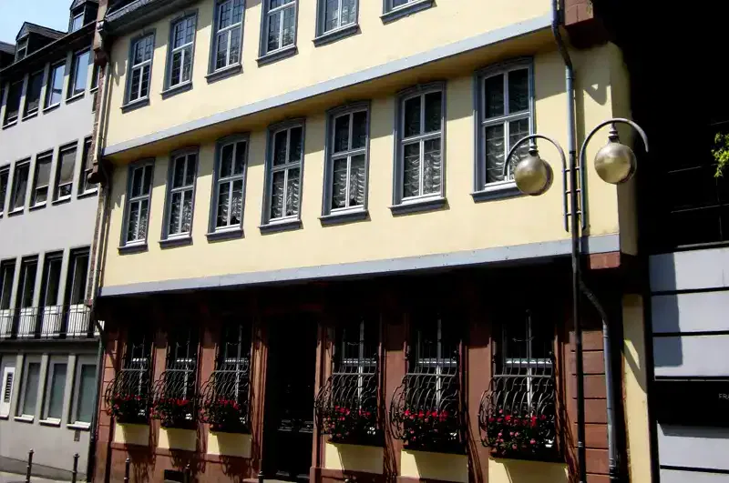Ein dreistöckiges Gebäude mit einer hellgelben Fassade und mehreren Fenstern, jedes mit weißen Vorhängen und Blumenkästen im unteren Stockwerk. Der Vintage-Look dieses Gebäudes, das an das Goethe-Haus erinnert, umfasst einen Laternenpfahl an der Vorderseite und angrenzende Gebäude, die auf beiden Seiten sichtbar sind.