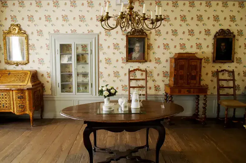 Ein Zimmer im Vintage-Stil mit Blumentapete, Holzboden und einem runden Holztisch mit weißen Keramikstücken. Der Raum erinnert an das Goethe-Haus und verfügt über antike Möbel wie einen Schrank, Stühle, einen Kronleuchter und Porträts an den Wänden, die den Charme der alten Welt ausstrahlen.