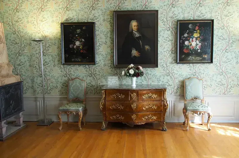 Ein aufwendig dekorierter Raum, der an das Goethe-Haus erinnert. Er verfügt über eine Holzkommode mit aufwendigen Goldverzierungen, flankiert von zwei kunstvoll geschnitzten Stühlen, die mit hellblauem Stoff bezogen sind. Über der Kommode hängen ein großes Porträt und zwei Blumengemälde. Die Wände sind mit Blumentapeten bedeckt.