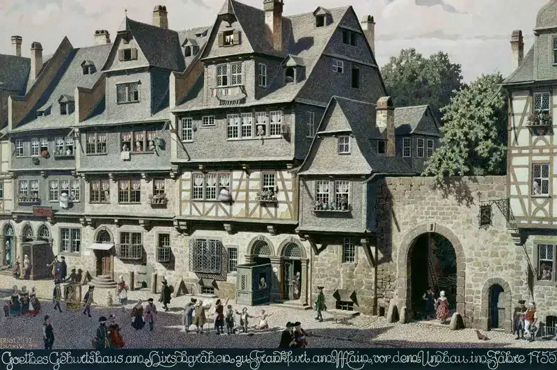 Eine geschäftige Straßenszene vor dem historischen Goethe-Haus, einem Fachwerkhaus. Menschen gehen spazieren und unterhalten sich, darunter auch spielende Kinder. Das Gebäude verfügt über mehrere Giebeldächer und einen Eingang mit einem Steinbogen. Im Hintergrund sind üppige grüne Bäume zu sehen.