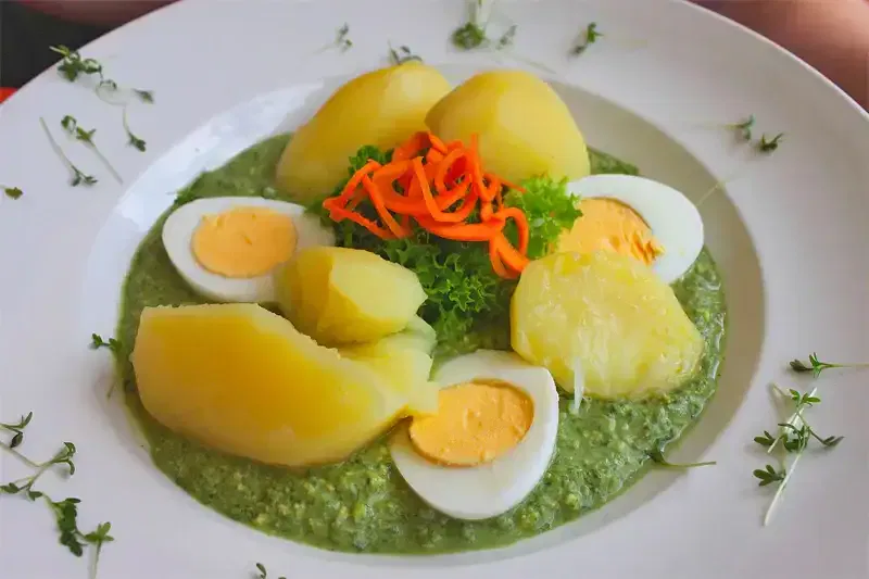 Ein Teller mit gekochten gelben Kartoffeln, geschnittenen hartgekochten Eiern und einer leuchtend grünen Soße. Das Gericht ist mit krauser Petersilie und dünnen Streifen roter Paprika garniert. Dünne grüne Garnituren sind um den Rand des weißen Tellers verstreut.