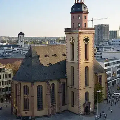 Ein hoher, brauner Glockenturm erhebt sich über einer großen, historischen Kirche mit einem detailreichen Dach, gelegen in einem Stadtgebiet mit modernen Gebäuden und Baukränen im Hintergrund. Dieses ikonische Bauwerk ist eine der bemerkenswertesten Sehenswürdigkeiten der Stadt.