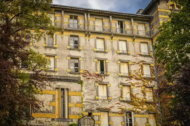 Ein verwittertes, mehrstöckiges Hotel, an dessen Wänden Efeu hochwächst, mit gelben Akzenten um die Fenster und Türen. Die alte Fassade des Gebäudes ist von üppigen Bäumen umgeben und der Himmel darüber ist teilweise bewölkt – eine einladende Szene im C&G Hotel.