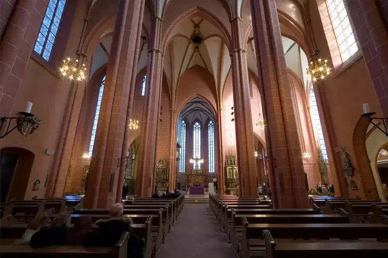 Ein Blick ins Innere des Kaiserdoms offenbart eine große Kathedrale mit hohen Gewölbedecken und großen Bogenfenstern. Reihen von Holzbänken säumen den Boden und erstrecken sich bis zum reich verzierten, mit goldenen Akzenten geschmückten Altar. Kronleuchter hängen von der Decke und einige Menschen sitzen in den Bänken.