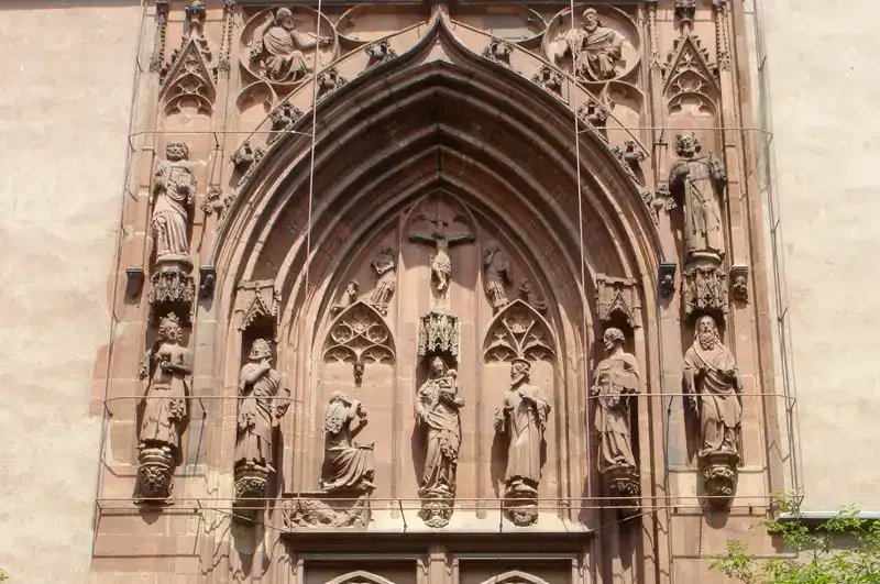 Eine kunstvolle Kirchenfassade im gotischen Stil mit detaillierten Schnitzereien von Statuen, Bögen und aufwendigem Mauerwerk. Der Eingang zu diesem Kaiserdom ist mit verschiedenen religiösen Figuren und Engeln in Nischen geschmückt, umgeben von dekorativen Elementen, die die architektonische Schönheit und Handwerkskunst betonen.