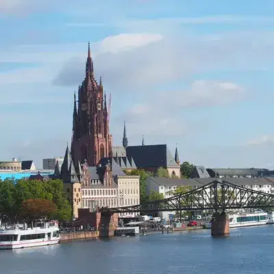 Blick auf Frankfurt, Deutschland, mit dem ikonischen Frankfurter Dom im gotischen Stil mit seinem hohen Turm, eingebettet zwischen historischen Gebäuden. Die Szene umfasst auch den Main mit Booten und einer Metallbrücke im Vordergrund unter einem klaren blauen Himmel, die einige der bemerkenswerten Sehenswürdigkeiten der Stadt zeigt.