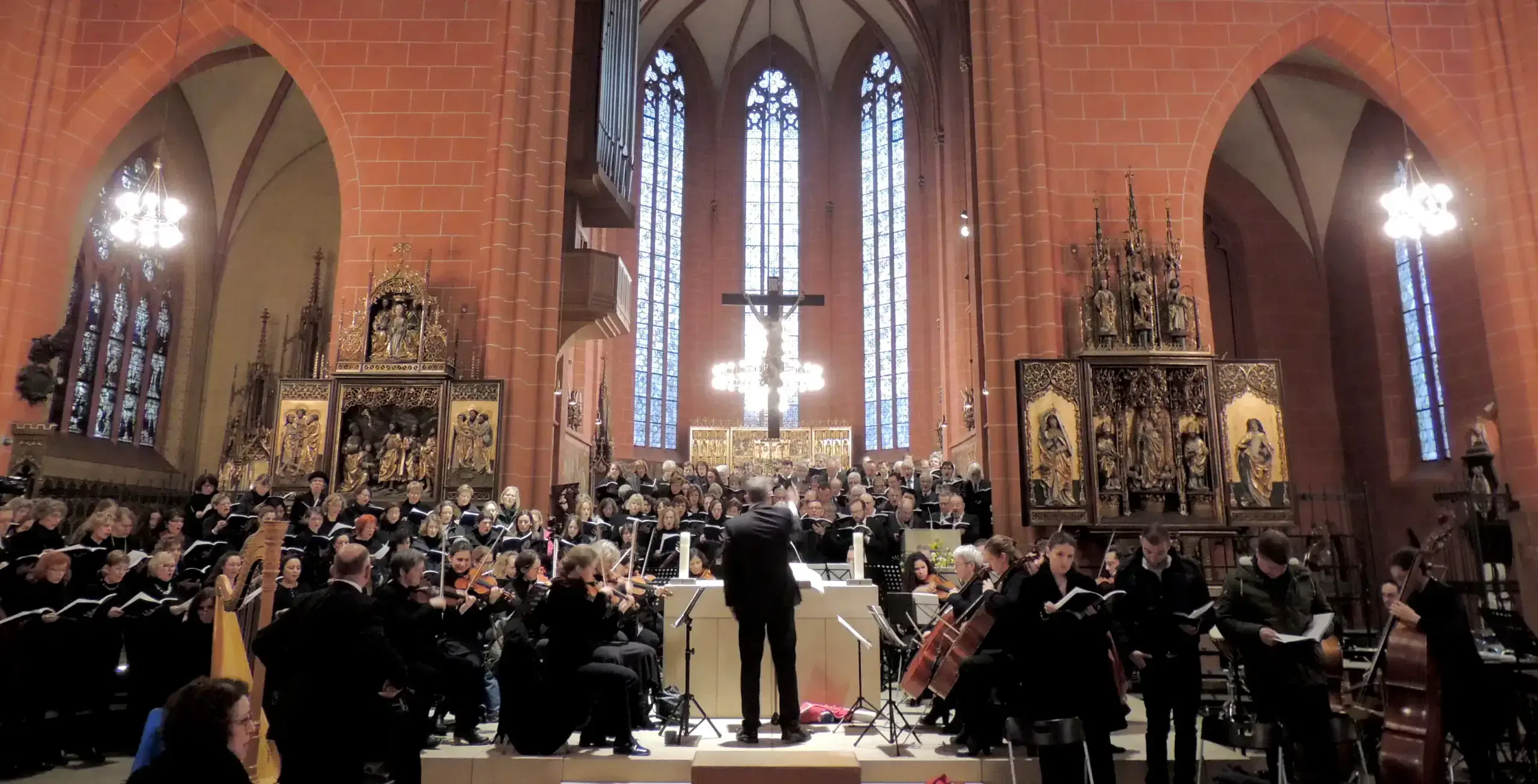 Ein Dirigent leitet ein Orchester und einen Chor, die in einem prachtvollen Kaiserdom mit hohen Gewölbedecken und Buntglasfenstern auftreten. Die Chormitglieder und Musiker sind in abgestuften Reihen auf einer erhöhten Plattform angeordnet, umgeben von kunstvollen Altarbildern und warmer Beleuchtung.