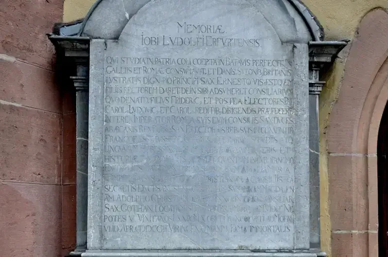 Eine alte Steintafel mit lateinischen Inschriften zu Ehren von Iob Ludovic Heupertensis. Der Text ist in den Stein gehauen, vor einer Backsteinmauer, die an die Architektur der Hauptwache erinnert, und wird von einer dekorativen Steinumrandung eingerahmt. Der Hintergrund enthält Teile einer gelben und roten Mauer.