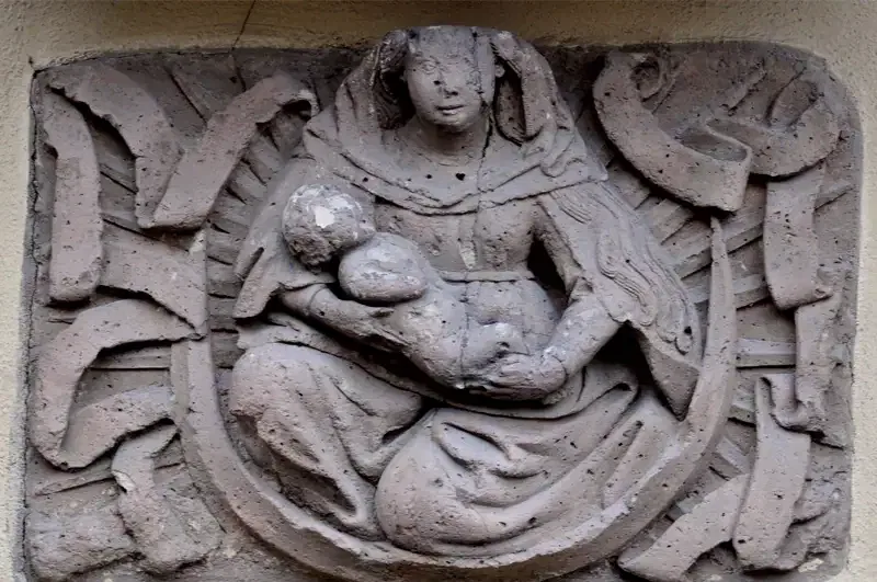 Ein Steinrelief an der Hauptwache zeigt eine sitzende Frau in mittelalterlicher Kleidung, die ein Kind in den Armen hält. Der Hintergrund weist komplizierte lineare Muster auf, die dem Kunstwerk Tiefe und Struktur verleihen. Die Figuren sind mit subtilen Details wiedergegeben, die ihre heiteren Ausdrücke betonen.
