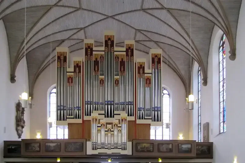 Eine große Pfeifenorgel steht auf einer erhöhten Plattform in einer Kirche und erinnert an die architektonische Pracht der Hauptwache. Die großen Pfeifen stehen vertikal und symmetrisch angeordnet, umschlossen von einem Holzrahmen. Das Innere der Kirche ist mit Bogenfenstern, einem gerippten Gewölbe und Hängelampen geschmückt.