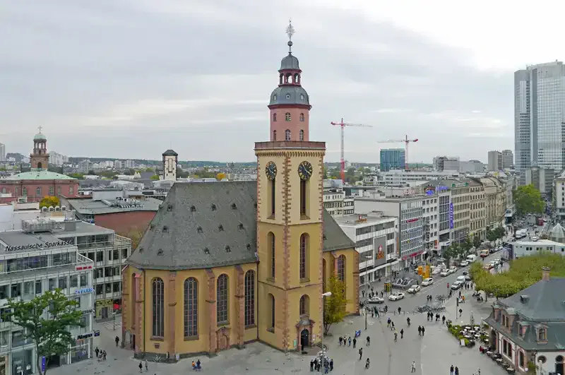 Eine Luftaufnahme der Katharinenkirche in der Nähe der Hauptwache in Frankfurt, Deutschland. Die historische Kirche mit ihrer unverwechselbaren Architektur sticht zwischen modernen Gebäuden und belebten Straßen hervor. Menschen haben sich um den Platz versammelt und im Hintergrund sind Kräne zu sehen.
