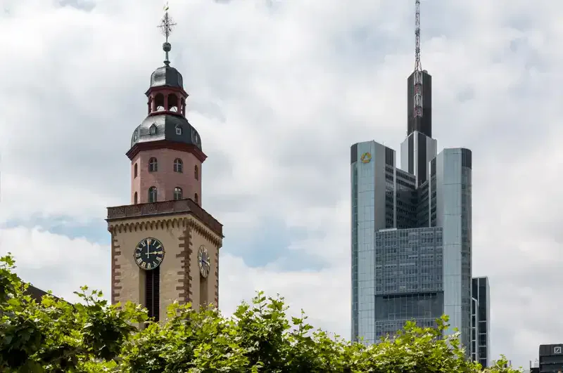 Das Bild zeigt zwei markante Gebäude in Frankfurt: die historische rote Backsteinkirche St. Katharina mit einem Uhrturm in der Nähe der geschäftigen Hauptwache und einen modernen Wolkenkratzer, den Commerzbank Tower, vor einem leicht bewölkten Himmel im Hintergrund. Im Vordergrund sind grüne Bäume zu sehen.