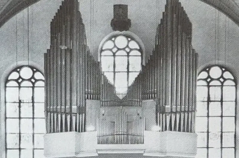 Schwarzweißfoto einer großen Pfeifenorgel vor der Hauptwachekirche mit hohen Bogenfenstern. Die Orgelpfeifen bilden ein symmetrisches Muster, das hoch zur Decke reicht und von komplizierten architektonischen Details eingerahmt wird.