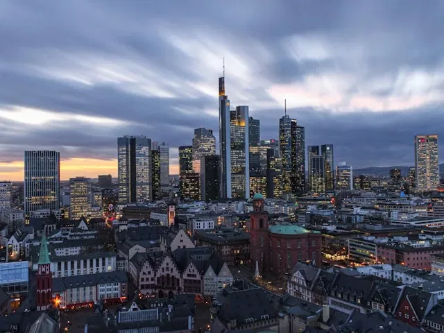 Eine Stadtsilhouette in der Abenddämmerung mit hohen modernen Wolkenkratzern und älteren traditionellen Gebäuden im Vordergrund, unter einem dramatischen, wolkenverhangenen Himmel mit einem Hauch von Sonnenuntergangslicht - perfekt, um Mobilität Frankfurt zu entdecken und Frankfurt ohne Auto zu erkunden.
