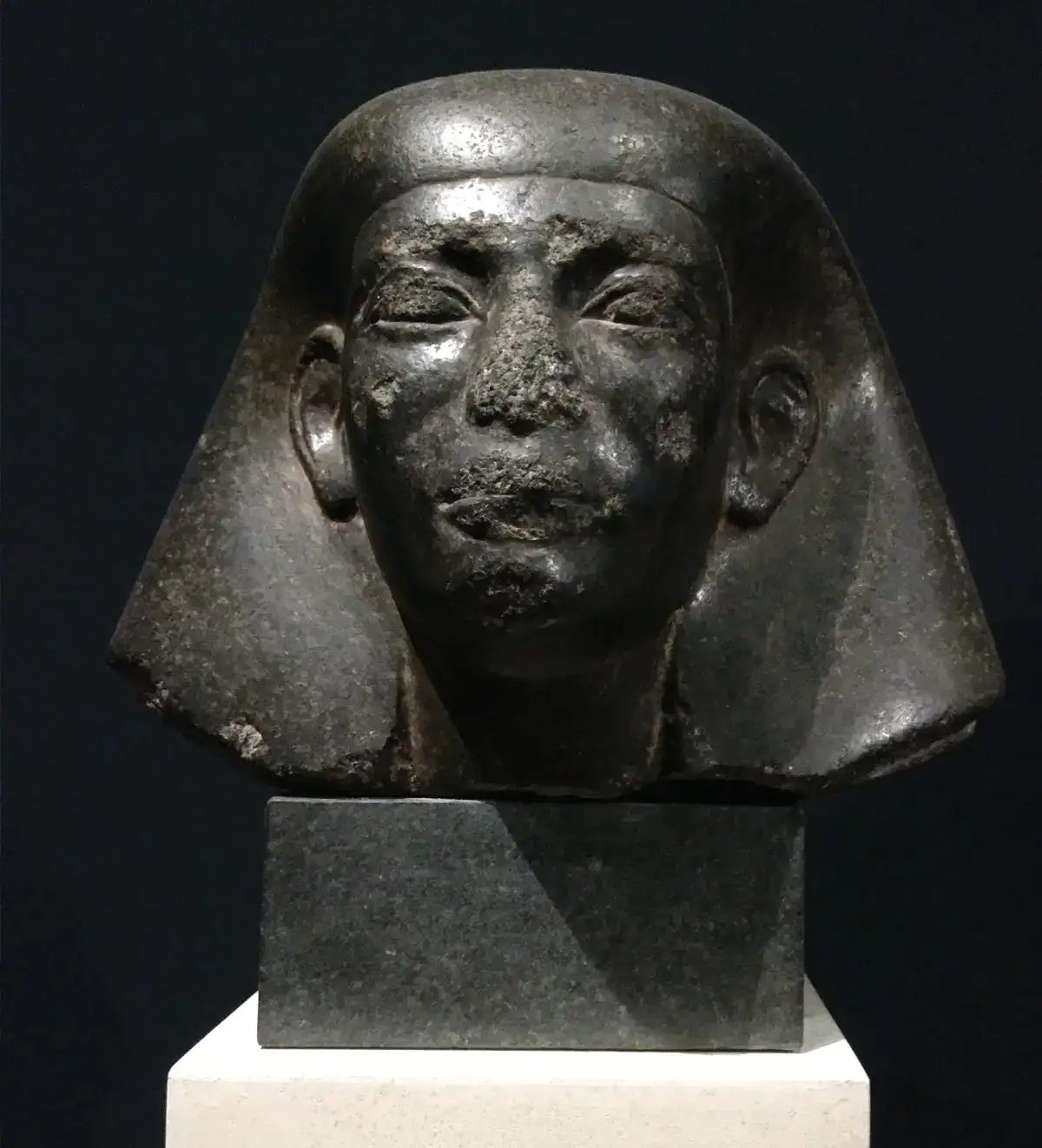 Eine schwarze Steinskulptur eines altägyptischen Kopfes mit einem glatten, rechteckigen Kopfschmuck, der das Gesicht umrahmt. Die Statue ist beschädigt, insbesondere um Nase und Mund herum, und wird während der Nacht der Museen auf einem weißen Sockel vor einem dunklen Hintergrund ausgestellt.