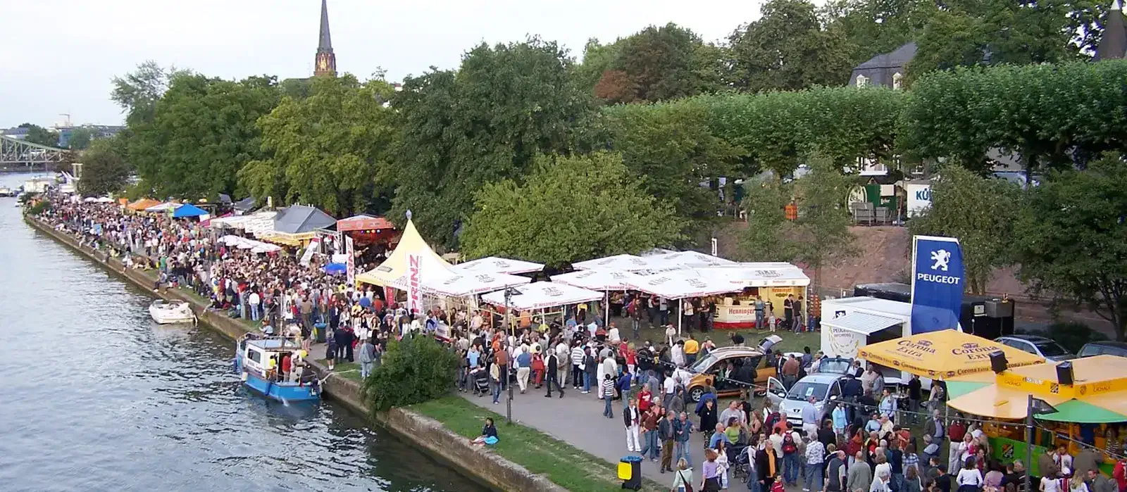 museumsuferfest-2005 Ein gut besuchtes Fest am Flussufer, das an das Museumsuferfest erinnert, bietet zahlreiche Zelte, Stände und Essensverkäufer. Die Menschen gehen den gepflasterten Weg entlang und genießen die lebhafte Atmosphäre. Im Hintergrund sind Bäume und ein Kirchturm zu sehen, die die malerische Kulisse noch verstärken.