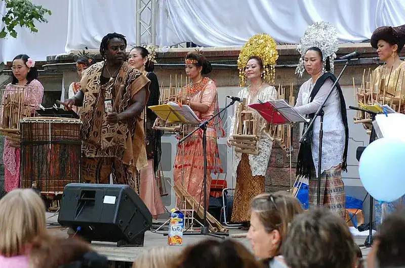 museumsuferfest-auftritt Eine bunt gemischte Gruppe von Musikern tritt während des Museumsuferfests auf einer Freilichtbühne für ein Publikum auf. Die Künstler, in farbenfrohe und traditionelle Kleidung aus verschiedenen Kulturen gekleidet, spielen verschiedene Musikinstrumente. Die Menge schaut aufmerksam zu, wobei einige Leute sitzen und andere stehen.