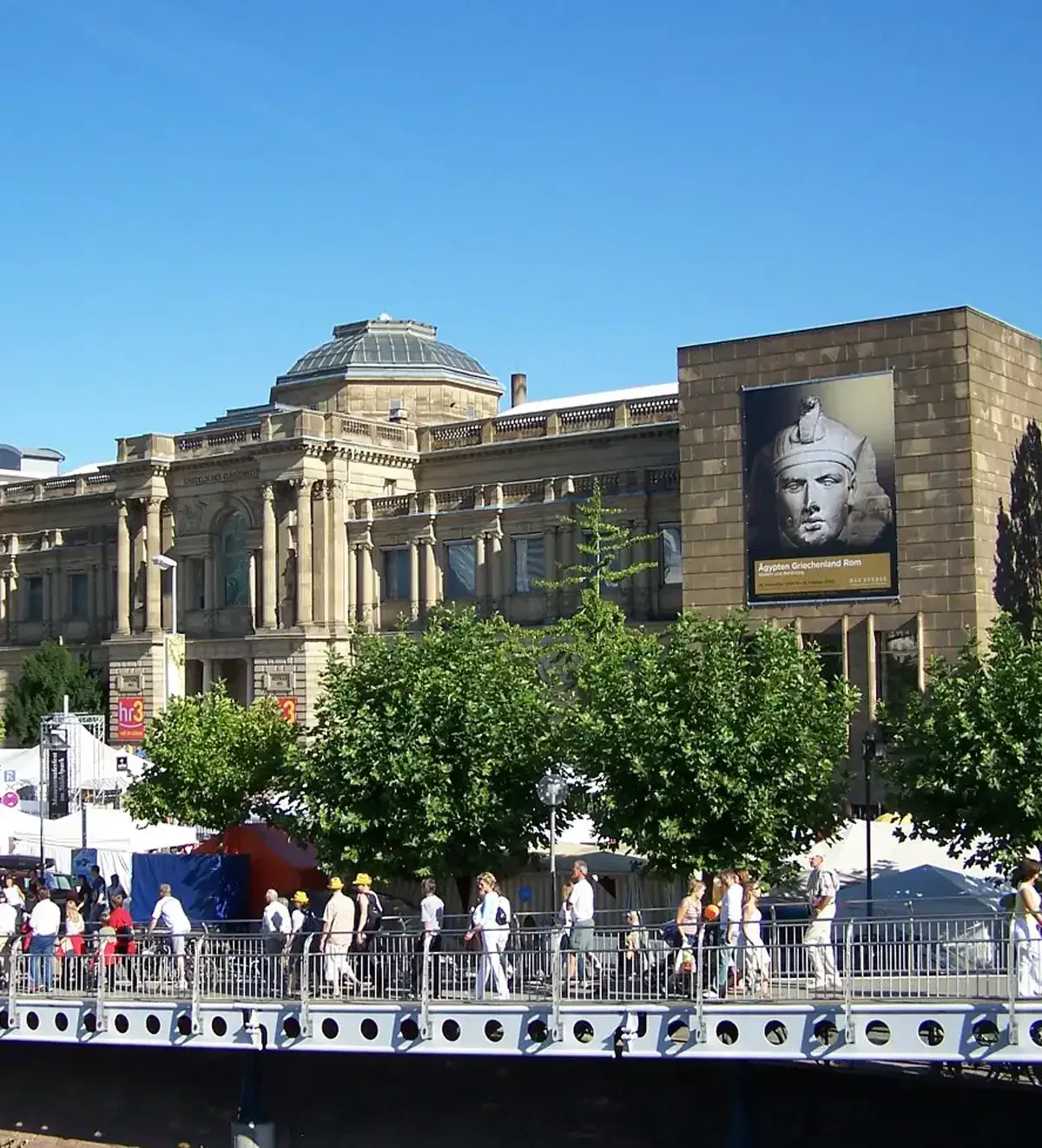 museumsuferfest-frankfurter-feste Im Hintergrund ist ein historisches Gebäude mit prachtvoller Architektur, Säulen und Kuppeldach zu sehen. Davor verläuft eine belebte Straße, auf der Fußgänger über eine Brücke laufen. Am Gebäude ist eine große Werbung für das Museumsuferfest angebracht, das eine Ausstellung mit einer antiken Statue präsentiert.