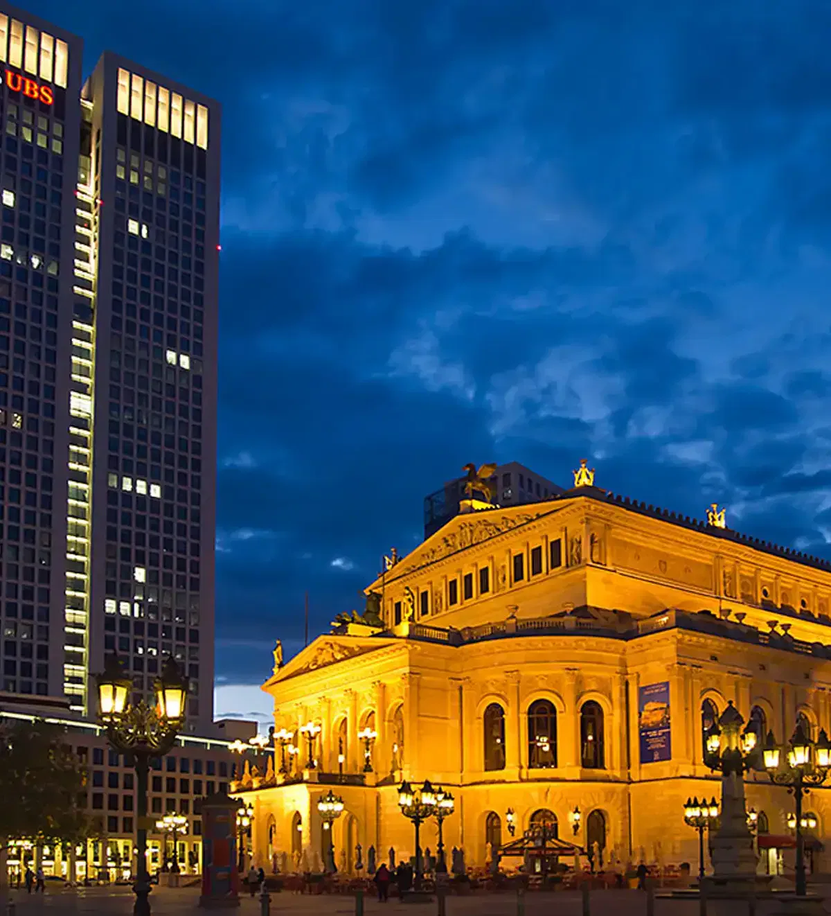 opernplatzfest-frankfurter-feste Ein großes, beleuchtetes Gebäude, wahrscheinlich ein historisches Opernhaus, ragt prominent unter einem Abendhimmel hervor. Im Hintergrund leuchtet ein hoher, moderner Wolkenkratzer mit dem „UBS“-Logo. Straßenlaternen und abendliche Lichter der Stadt heben die Szene hervor und fangen die lebendige Atmosphäre des Opernplatzfests ein.