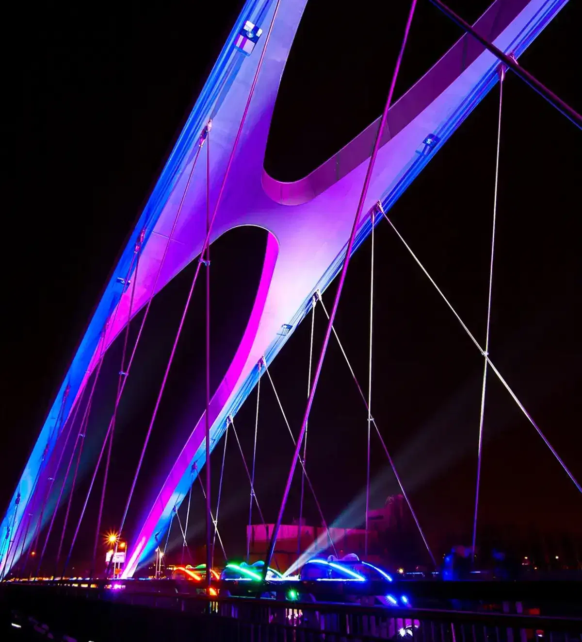 osthafen-festival-frankfurter-feste Eine moderne Brücke, die nachts mit leuchtenden Neonlichtern in Lila- und Rosatönen beleuchtet wird. Das elegante Design zeichnet sich durch deutlich geschwungene Bögen und mehrere Hängekabel aus, die eine auffällige, futuristische Ästhetik erzeugen. Während des Osthafenfestivals verleihen die Lichter darunter der Szene farbenfrohe Akzente.