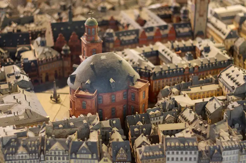 paulskirche-altstadtmodell Ein detailliertes Miniaturmodell einer historischen europäischen Stadt mit dicht gedrängten Gebäuden mit grauen Dächern und mehreren bemerkenswerten Bauwerken, darunter die Paulskirche und ein markantes Kuppelgebäude in der Mitte. Das Modell zeigt komplizierte architektonische Details und Stadtplanung.