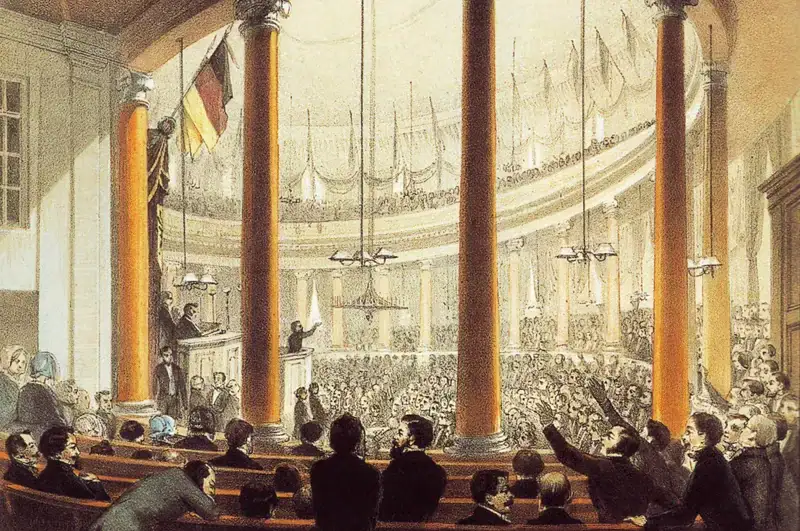 paulskirche-bilderrevolution Eine historische Illustration zeigt eine große, überfüllte Versammlung im großen Saal der Paulskirche mit hohen Säulen und hoher Decke. Eine Person am Rednerpult scheint zum Publikum zu sprechen, während die Anwesenden, einige gestikulierend, aufmerksam zuhören. Darüber hängen europäische Flaggen.