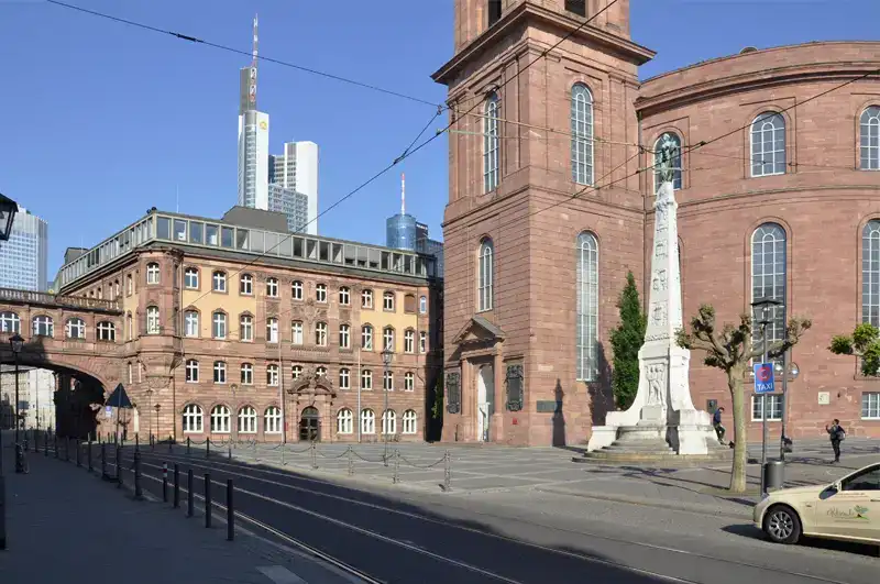 paulskirche-paulsplatz-mit-einheitsdenkmal Eine Stadtstraße in Frankfurt, Deutschland, zeichnet sich durch markante Backsteinarchitektur und eine hohe, vertikale Statue aus. Die Paulskirche steht stolz zwischen den historischen Gebäuden im Vordergrund, während im Hintergrund moderne Wolkenkratzer aufragen. Auf der Straße ist ein Taxi und wenig Verkehr.