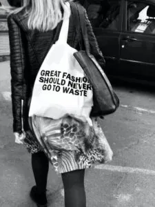 Schwarzweißfoto einer Person, die durch Frankfurt am Main läuft. Sie trägt eine Lederjacke, einen gemusterten Rock und eine Einkaufstasche mit dem Text „GROSSE MODE SOLLTE NIE VERSCHWENDEN“. Es ist ein ikonischer Einblick in den urbanen Einkaufsstil.