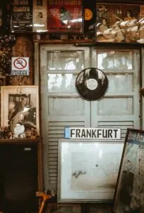 Ein Raum ist mit einer Vielzahl von Vintage-Artikeln, Schallplatten, Postern und einem Nummernschild mit der Aufschrift „Frankfurt am Main“ dekoriert. An den hellblauen Doppeltüren im hinteren Bereich ist eine Vinyl-Schallplatte angebracht, und an einem Schrank lehnen gerahmte Bilder, die den Charme klassischer Shopping-Möglichkeiten hervorrufen.