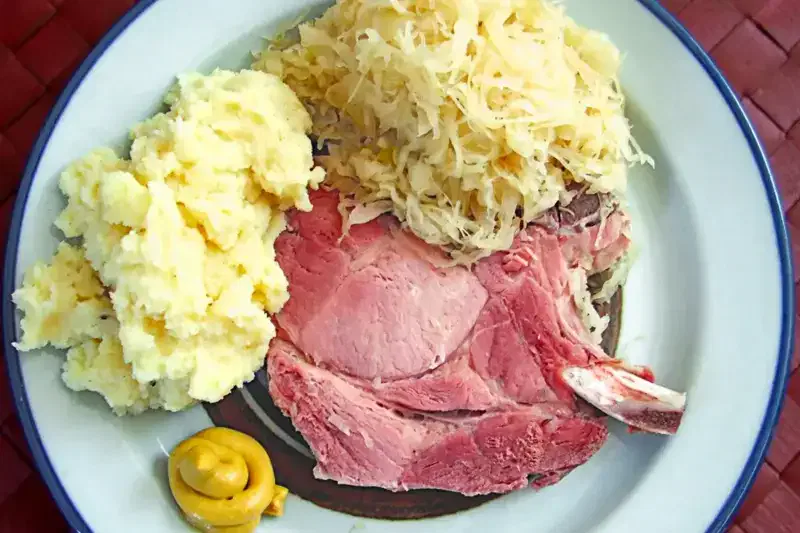 Ein Teller mit einer Portion Kartoffelbrei, einer Portion Sauerkraut und einer Scheibe gekochtem Schinken. Am Rand des Tellers befindet sich ein kleiner Klecks Senf.