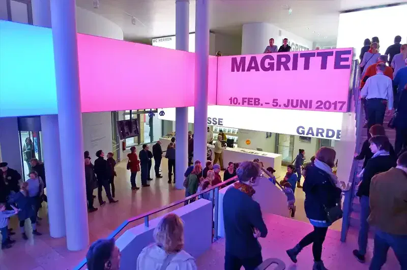 Ein belebter Innenraum mit versammelten Menschen. Ein großes Schild mit der Aufschrift „Magritte 10. Feb. – 5. Juni 2017“ ziert die Kunsthalle Schirn. Rosa und blaues Licht illuminieren die modernen Designelemente mit weißen Säulen und Glasfenstern.