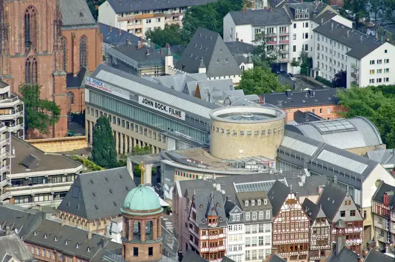 Luftaufnahme einer historischen europäischen Stadt mit einer Mischung aus alter und moderner Architektur. Zu den markanten Gebäuden gehören eine große Kirche mit einem hohen Kirchturm, traditionelle Gebäude mit schrägen Dächern und moderne Gebäude. Besonders hervorzuheben ist die Kunsthalle Schirn neben einem Banner mit der Aufschrift „JOHN BOCK FILME“.