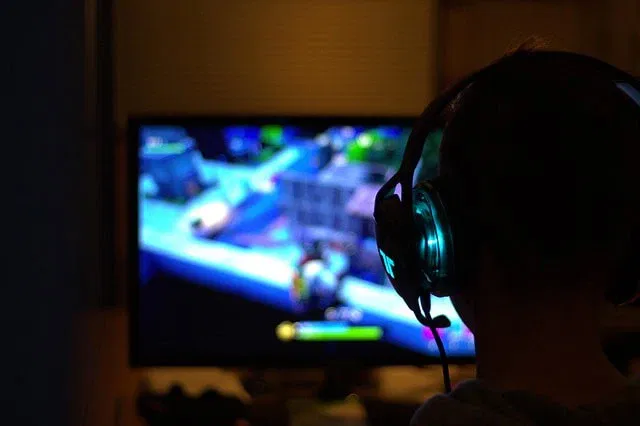 Eine Person mit großen Kopfhörern sitzt vor einem hell erleuchteten Computerbildschirm und spielt ein buntes Videospiel. Als einer der bekannten Deutschen Streamer genießt sie eine digitale Landschaft mit Gebäuden und Objekten in einem schwach beleuchteten Raum.