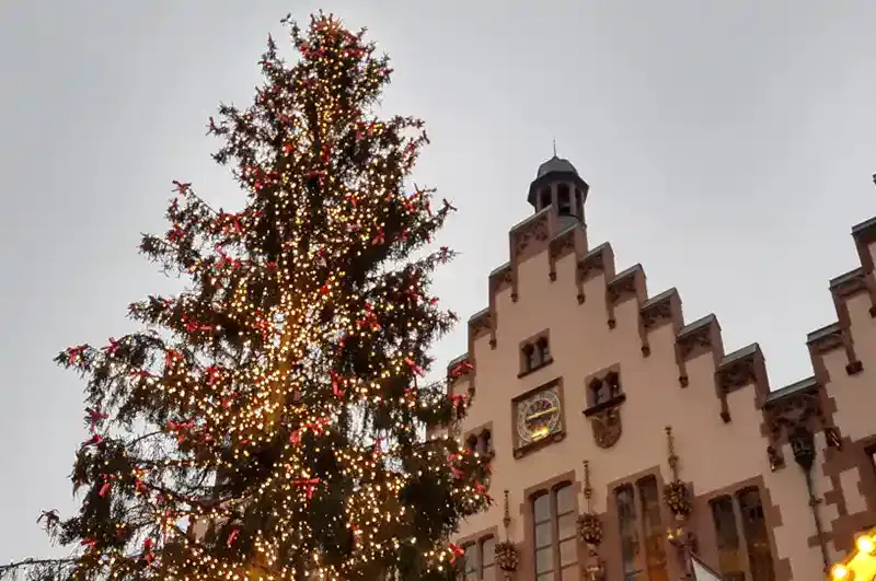 Ein großer, mit Lichterketten geschmückter Weihnachtsbaum steht vor einem historischen Gebäude mit Stufengiebeldach und Glockenturm und erinnert vor einem bewölkten Himmel an die festliche Stimmung eines traditionellen Weihnachtsmarkts.