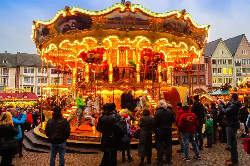 Ein hell erleuchtetes Karussell mit farbenfrohen Dekorationen und sich bewegenden Pferden ist von einer Menschenmenge umgeben, sowohl Kindern als auch Erwachsenen, die in der Abenddämmerung die festliche Atmosphäre auf einem Stadtplatz genießen. Der Hintergrund, der an einen Weihnachtsmarkt erinnert, zeigt farbenfrohe Gebäude und funkelnde Lichter.