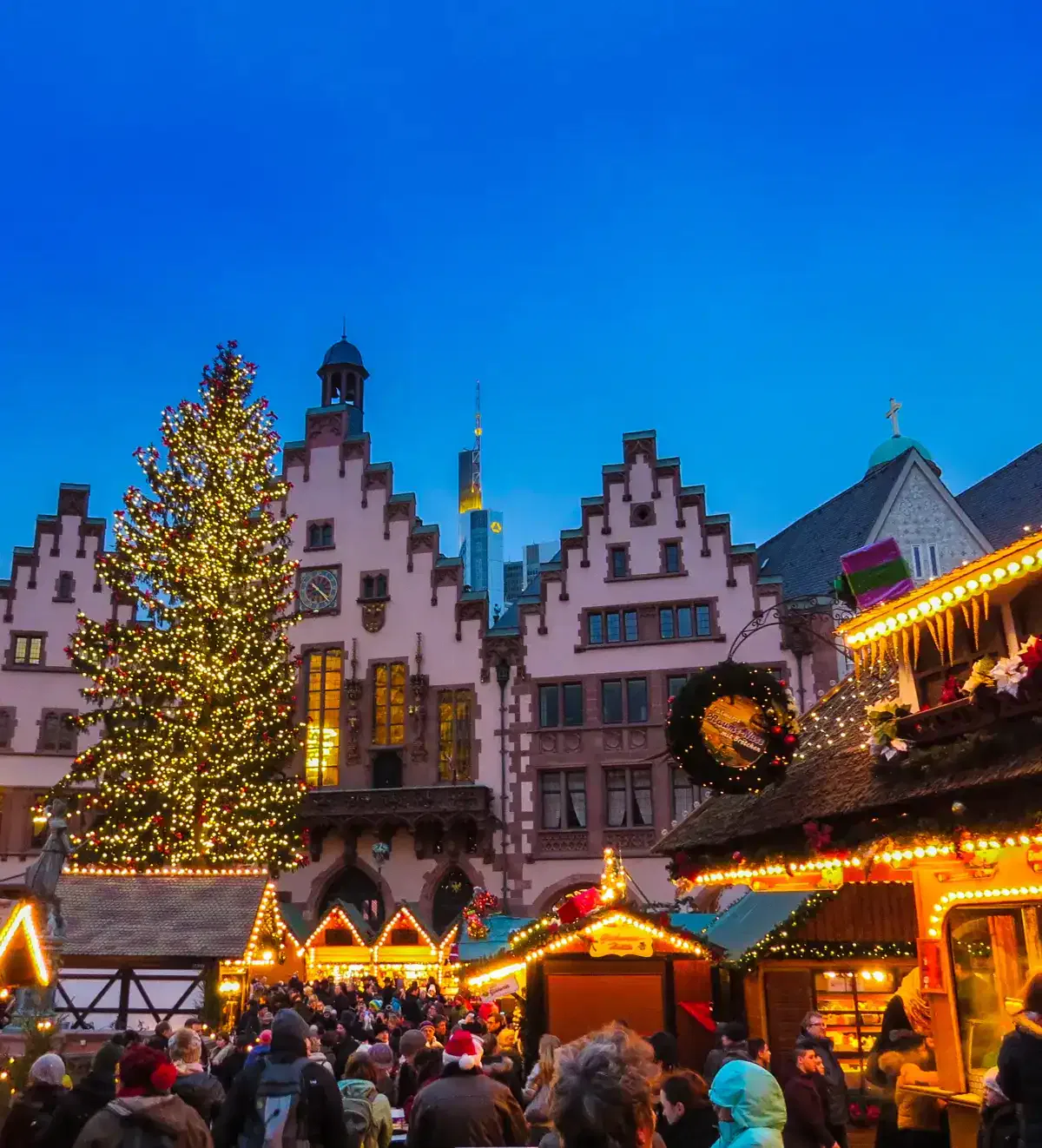 Abends versammeln sich Menschenmassen auf einem festlichen Weihnachtsmarkt. Die Szene ist mit funkelnden Lichtern, dekorierten Ständen und einem großen beleuchteten Weihnachtsbaum geschmückt. Historische Giebelhäuser bilden eine malerische Kulisse unter einem klaren, dunkelblauen Himmel.
