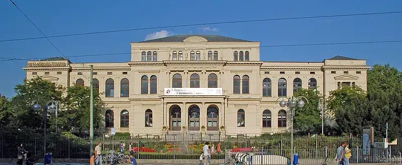 In der Nähe des Frankfurter Zoos steht ein großes, historisches, beiges Gebäude mit drei Stockwerken und Bogenfenstern. Der Mittelteil verfügt über einen markanten Eingang mit Treppe. An der Vorderseite hängt ein Banner, und die Straße davor ist von Menschen zu Fuß oder mit dem Fahrrad befahren. Der Himmel ist klar und blau.