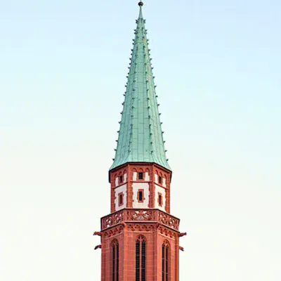 Der hohe Turm der Alten Nikolaikirche mit seinem grünen Spitzdach bildet einen Kontrast zum klaren, hellblauen Himmel. Der Kirchturm ist Teil einer roten Backsteinkonstruktion mit weißen architektonischen Details um die Dachbasis.