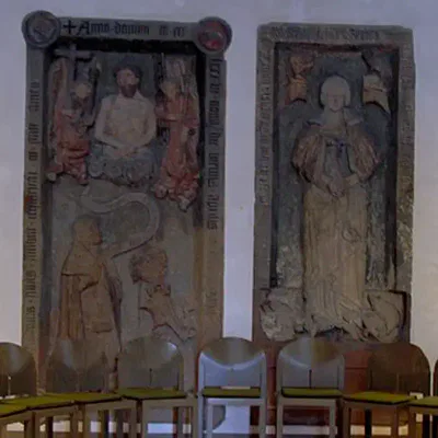 Ein Foto, das zwei alte Grabsteingravuren zeigt, die an einer weißen Wand in der historischen Alten Nikolaikirche angebracht sind. Die linke Gravur zeigt eine große zentrale Figur mit kleineren Figuren darunter, während die rechte Gravur eine einzelne stehende Figur zeigt. Vor den Gravuren stehen fünf Stühle aufgereiht.