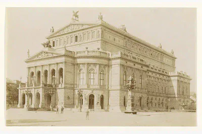 Ein Sepiafoto zeigt die Alte Oper, ein kunstvolles, mehrstöckiges klassisches Gebäude mit detaillierten architektonischen Elementen, darunter Statuen, Säulen und Bogenfenster. Es scheint aus einer historischen Epoche zu stammen. Auf dem Platz davor sind ein paar Menschen und ein großer, kunstvoll verzierter Laternenpfahl.