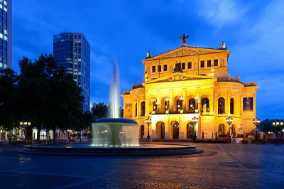Das Bild zeigt die beleuchtete Alte Oper in Frankfurt in der Abenddämmerung. Das historische Gebäude zeichnet sich durch eine großartige Architektur mit Bogenfenstern und Säulen aus. Im Vordergrund steht ein beleuchteter Brunnen, im Hintergrund sind moderne Gebäude zu sehen.