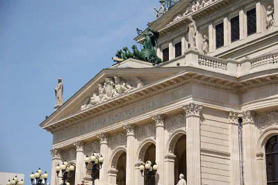 Das Foto zeigt die kunstvolle Fassade der Alten Oper, einem Konzertsaal in Frankfurt, Deutschland. Die Architektur zeichnet sich durch klassische Säulen, Statuen und detailreiche Reliefs aus. Die Aufschrift „DEM WAHREN SCHÖNEN GUTEN“ ist unter der Dachlinie vor einem klaren blauen Himmel sichtbar.