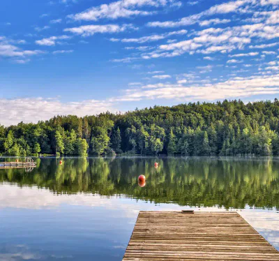 Ein Holzsteg erstreckt sich in einen ruhigen See, der von üppigen grünen Bäumen umgeben ist. Das ruhige Wasser spiegelt den Wald und den teilweise bewölkten Himmel darüber wider. Mehrere orangefarbene Bojen schwimmen auf dem Wasser und verleihen der friedlichen Szene einen Hauch von Farbe.