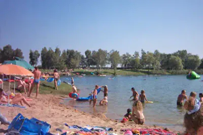 Eine überfüllte Strandszene an einem See an einem sonnigen Tag. Menschen schwimmen, faulenzen unter bunten Sonnenschirmen und Kinder spielen im Sand. Im Hintergrund säumen Bäume das Ufer und im Wasser sind aufblasbare Spielzeuge zu sehen.
