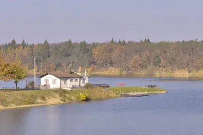 Eine ruhige Seeszene mit einem kleinen Haus auf einer schmalen Halbinsel, umgeben von Wasser. Das Ufer ist von Bäumen in herbstlichen Farben gesäumt. In der Nähe liegt ein rotes Boot vor Anker, und der Himmel ist klar und blau, was auf einen ruhigen, friedlichen Tag schließen lässt.