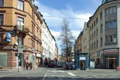Eine belebte Stadtstraße mit mehrstöckigen Gebäuden, in denen sich im Erdgeschoss Geschäfte und darüber Wohneinheiten befinden, erwacht während des Festes zum Leben. Menschen gehen auf den Bürgersteigen, während entlang der Straße Autos geparkt sind. Ein strahlend blauer Himmel mit vereinzelten Wolken bildet die festliche Kulisse.